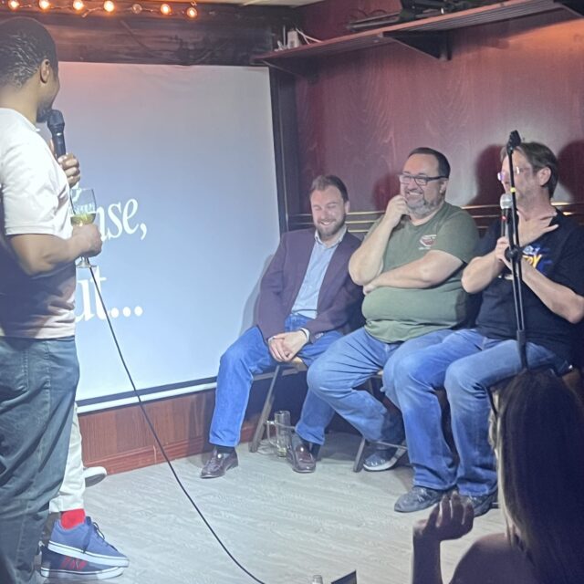 Comedian on stage with three guests seated in front of the No Offense, But... screen