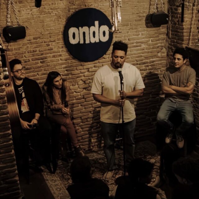 Comedian performing in a brick-walled room at a No Offense, But... show