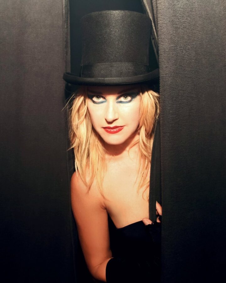 Rachel Arieff in a black top hat, looking out from between stage curtains